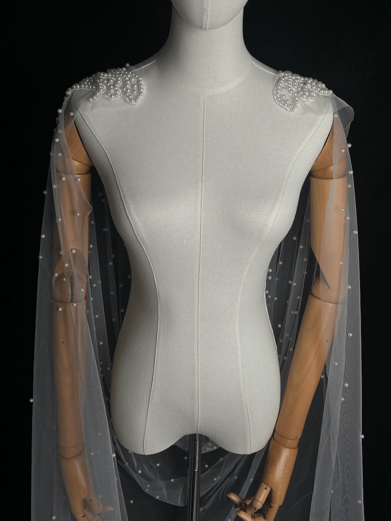 May include: A sheer white tulle capelet featuring scattered white pearls throughout the fabric. The shoulders are adorned with dense, decorative clusters of white pearls arranged in a floral-like pattern, extending into long, flowing panels.