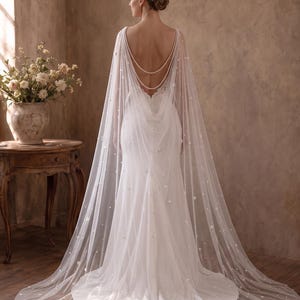 May include: A floor-length white wedding dress featuring a sheer, flowing cape overlay adorned with scattered pearls and small floral appliqu&eacute;s. The cape is draped over the shoulders and back, accented by two delicate strands of white pearls.