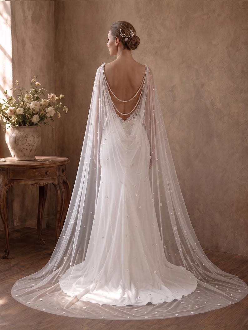 May include: A floor-length white wedding dress featuring a sheer, flowing cape overlay adorned with scattered pearls and small floral appliqu&eacute;s. The cape is draped over the shoulders and back, accented by two delicate strands of white pearls.