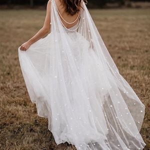 May include: A white floor-length gown featuring a sheer tulle cape overlay adorned with small white floral appliqu&eacute;s. The back of the dress showcases a deep scoop neckline accented by two draped strands of white pearls that connect at the shoulders.