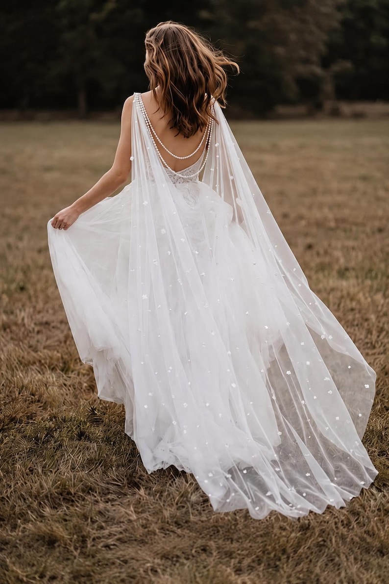 May include: A white floor-length gown featuring a sheer tulle cape overlay adorned with small white floral appliqu&eacute;s. The back of the dress showcases a deep scoop neckline accented by two draped strands of white pearls that connect at the shoulders.
