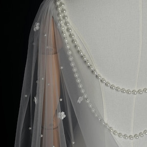 May include: A white sheer tulle capelet featuring delicate white floral appliqu&eacute;s and scattered faux pearls. The garment is adorned with two draped strands of white pearls that extend from the shoulder, creating a layered effect over the fabric.