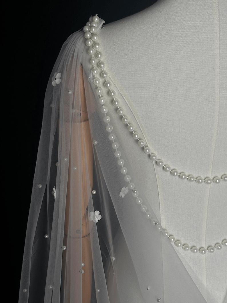 May include: A white sheer tulle capelet featuring delicate white floral appliqu&eacute;s and scattered faux pearls. The garment is adorned with two draped strands of white pearls that extend from the shoulder, creating a layered effect over the fabric.