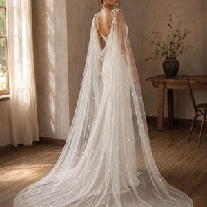May include: A floor-length white bridal gown featuring a sleeveless bodice with a deep back and a long, sheer tulle cape attached at the shoulders. The cape is adorned with scattered white pearls and extends into a sweeping train on the floor.