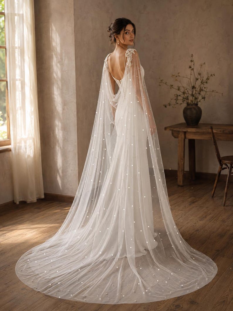 May include: A floor-length white bridal gown featuring a sleeveless bodice with a deep back and a long, sheer tulle cape attached at the shoulders. The cape is adorned with scattered white pearls and extends into a sweeping train on the floor.