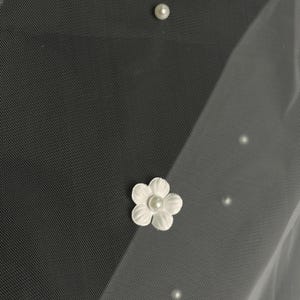 May include: A sheer white veil fabric featuring scattered small round white pearls and a single white fabric flower applique with a central pearl accent. The material has a fine mesh texture and is layered to create a soft, translucent appearance.