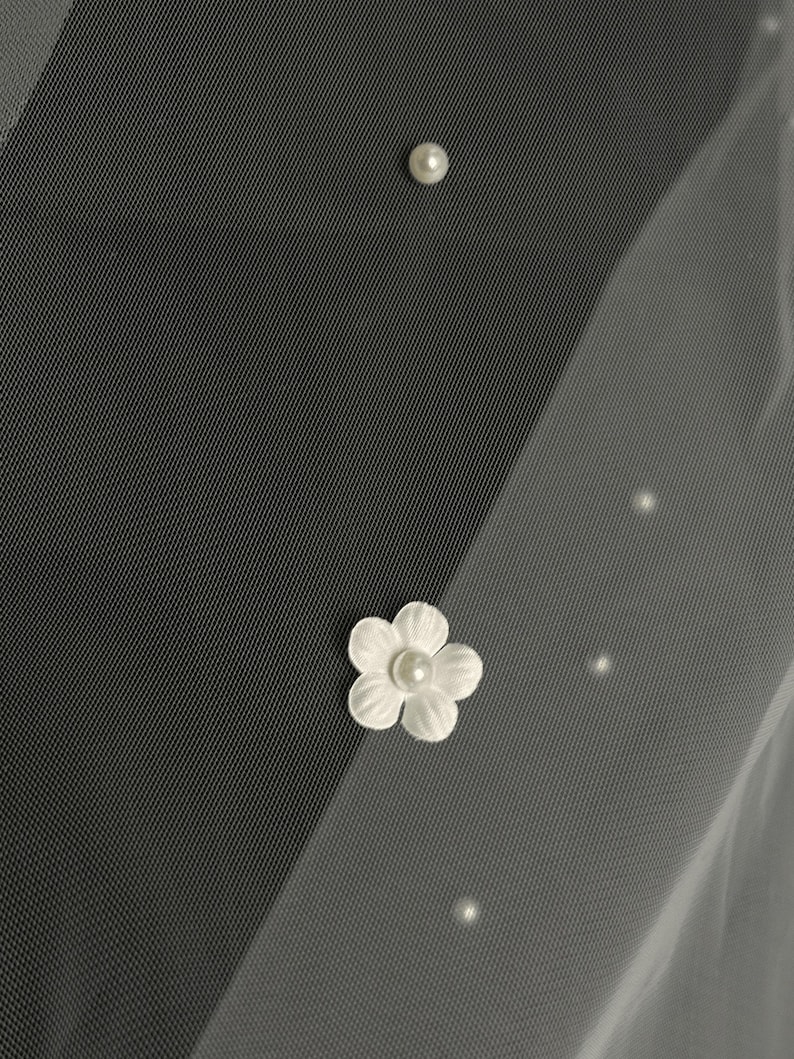 May include: A sheer white veil fabric featuring scattered small round white pearls and a single white fabric flower applique with a central pearl accent. The material has a fine mesh texture and is layered to create a soft, translucent appearance.