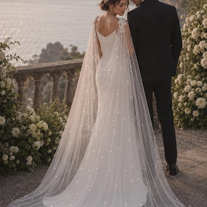 May include: A floor-length white wedding dress with a V-neckline and a long, sheer, floor-sweeping cape featuring delicate white floral lace appliques at the shoulders and scattered pearl-like embellishments throughout the fabric.