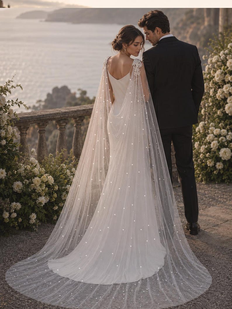 May include: A floor-length white wedding dress with a V-neckline and a long, sheer, floor-sweeping cape featuring delicate white floral lace appliques at the shoulders and scattered pearl-like embellishments throughout the fabric.