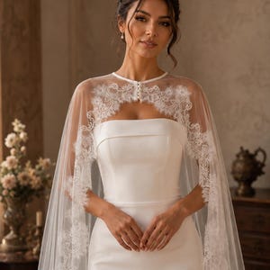 May include: A white strapless gown paired with a sheer, floor-length tulle cape featuring intricate white lace trim along the edges. The cape fastens at the neck with a row of small, round white buttons, creating a layered, elegant bridal ensemble.