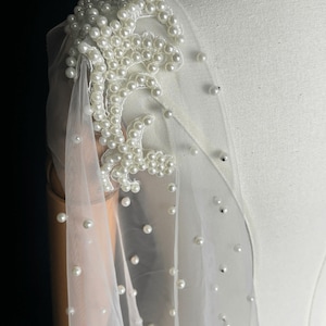 May include: A sheer white tulle fabric shoulder wrap featuring an intricate floral lace appliqu&eacute; densely embellished with round white pearls. The lightweight material is scattered with individual pearls throughout, creating a delicate texture.
