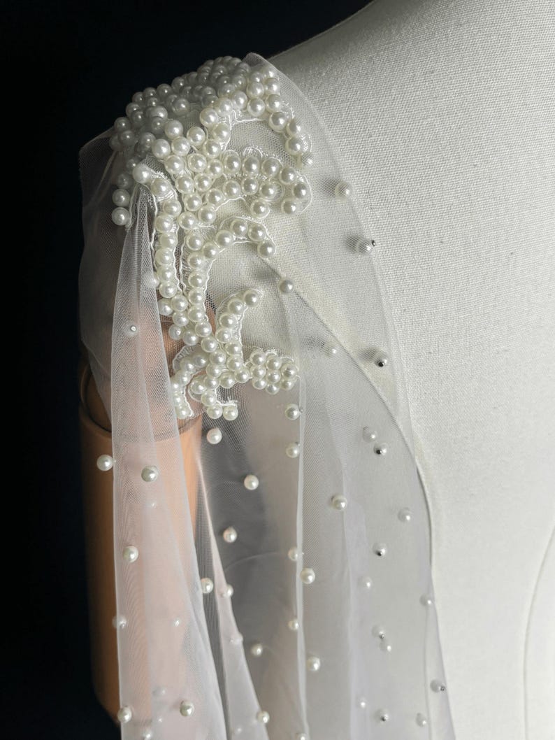 May include: A sheer white tulle fabric shoulder wrap featuring an intricate floral lace appliqu&eacute; densely embellished with round white pearls. The lightweight material is scattered with individual pearls throughout, creating a delicate texture.