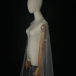 May include: A sheer white floor-length cape made of tulle fabric, featuring delicate white floral appliqu&eacute;s and scattered small white pearls. The cape attaches at the shoulders with a cluster of larger pearls and drapes down the back.