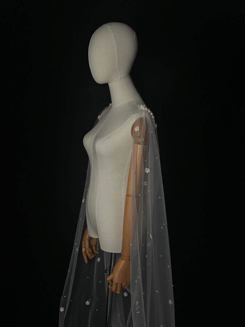 May include: A sheer white floor-length cape made of tulle fabric, featuring delicate white floral appliqu&eacute;s and scattered small white pearls. The cape attaches at the shoulders with a cluster of larger pearls and drapes down the back.