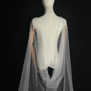 May include: A floor-length white tulle cape featuring dense clusters of white pearls on the shoulders and scattered individual pearls throughout the sheer fabric. The cape attaches at the shoulders and drapes down the back in a flowing silhouette.