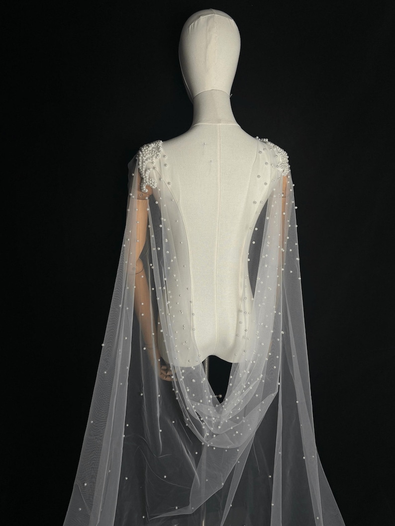 May include: A floor-length white tulle cape featuring dense clusters of white pearls on the shoulders and scattered individual pearls throughout the sheer fabric. The cape attaches at the shoulders and drapes down the back in a flowing silhouette.