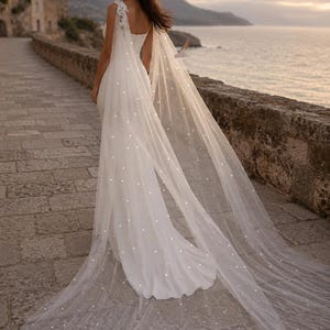 May include: A floor-length white gown featuring a fitted bodice and a long, sheer tulle cape attached at the shoulders. The cape is adorned with scattered white pearls and features delicate lace detailing at the shoulder attachment points.
