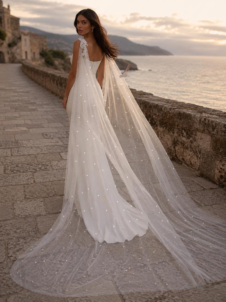 May include: A floor-length white gown featuring a fitted bodice and a long, sheer tulle cape attached at the shoulders. The cape is adorned with scattered white pearls and features delicate lace detailing at the shoulder attachment points.