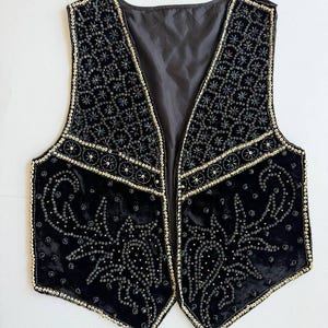 May include: A black velvet waistcoat featuring intricate beadwork and sequin embellishments. The garment displays a detailed pattern of floral and paisley motifs in black and gold tones, with a solid black lining and a sleeveless, open-front design.