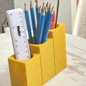 May include: A yellow three-tiered desk organiser with a geometric diamond pattern on the exterior. It holds several blue pencils, a red pen, a clear pen, and a white Helix ruler with markings up to 9 cm, showing the scale of the container.