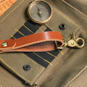 May include: A brown leather keychain featuring a brass key ring on one end and a brass swivel lobster clasp on the other. The leather strap is secured with a brass rivet and is stamped with the text Drake Adventure Supply on the inner side.