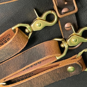 May include: Brown leather straps featuring brass swivel lobster clasps and metal rivets. The leather is stamped with the text DRAKE ADVENTURE SUPPLY. The straps include a circular metal key ring and a rectangular buckle with a copper-toned finish.