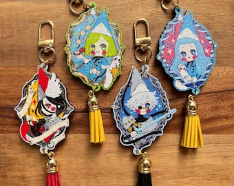 May include: Four decorative keychains featuring illustrated anime-style characters in pointed hats, each framed in a unique shape with a gold-tone metal clasp and a dangling suede tassel in red, yellow, black, or yellow, showcasing vibrant colors.