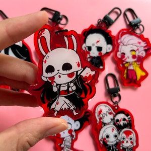 May include: A set of six red and black acrylic keychains featuring various horror characters. Each keychain has a black metal ring and a clear acrylic charm with a cartoon character design. The characters include a bunny rabbit with a bloody axe, a man with a bloody face, a man with a bloody face and a yellow jacket, a man with a bloody face and a black jacket, a man with a bloody face and a black jacket, and a man with a bloody face and a black jacket.