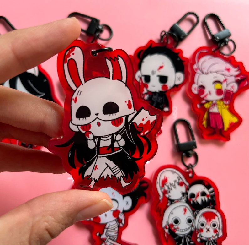 May include: A set of six red and black acrylic keychains featuring various horror characters. Each keychain has a black metal ring and a clear acrylic charm with a cartoon character design. The characters include a bunny rabbit with a bloody axe, a man with a bloody face, a man with a bloody face and a yellow jacket, a man with a bloody face and a black jacket, a man with a bloody face and a black jacket, and a man with a bloody face and a black jacket.