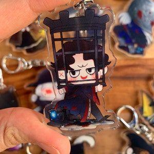 K&ouml;nnte beinhalten: Ein durchsichtiger Acryl-Schl&uuml;sselanh&auml;nger mit einer Comicfigur mit schwarzen Haaren, einem rot-schwarzen Outfit und einem K&auml;fig &uuml;ber dem Kopf. Die Figur h&auml;lt den K&auml;fig mit beiden H&auml;nden und hat einen schelmischen Gesichtsausdruck. Der Schl&uuml;sselanh&auml;nger ist von anderen Schl&uuml;sselanh&auml;ngern mit verschiedenen Comicfiguren umgeben.