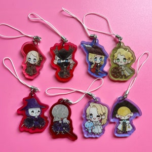 May include: A collection of eight acrylic charms with cartoon character designs. Each charm features a unique character with detailed outfits and expressions, attached to a white string. The charms are in various colors, set against a pink background.