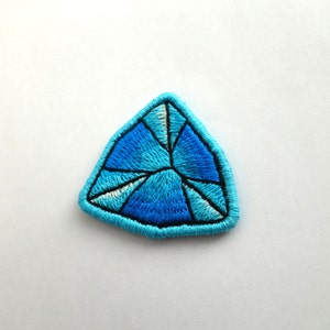 Pode incluir: Um remendo bordado azul em forma de diamante. O diamante é contornado com fio preto e tem um desenho de estrela branca dentro.