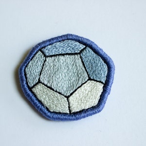 Può includere: Una toppa geometrica ricamata blu e bianca a forma di pietra preziosa. La toppa è realizzata in tessuto e ha un contorno cucito.