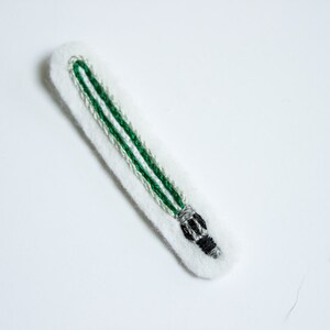 May include: A green lightsaber embroidered on white felt. The lightsaber has a black handle and a silver tip.