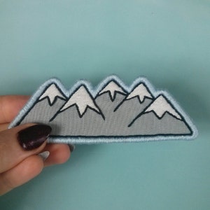 May include: A gray felt mountain range patch with white embroidered snow peaks. The patch is outlined in blue thread.