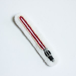 Può includere: Una toppa ricamata a forma di spada laser rossa e bianca con un'impugnatura nera.