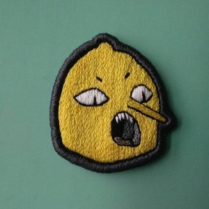 Puede incluir: Parche bordado con un personaje amarillo con forma de limón y contorno negro. El personaje tiene ojos blancos, una nariz larga y una boca abierta con dientes. El parche está sobre un fondo turquesa.