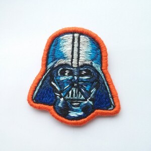 May include: Embroidered patch of Darth Vader's face with a blue and white helmet and black mask. The patch is outlined in orange thread.