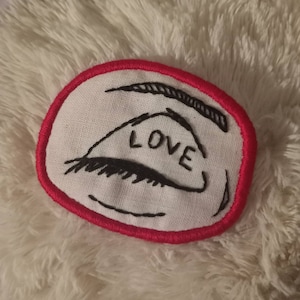 May include: A white fabric patch with a red border, featuring a black embroidered eye with the word "LOVE" written inside.