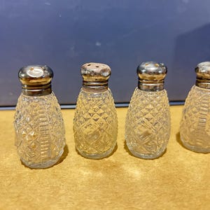 May include: Four small, clear glass shakers with a diamond-cut pattern and metallic silver-toned caps. Each shaker features a rounded, tapered body with a textured surface and a smooth, reflective metal lid with small holes for dispensing contents.