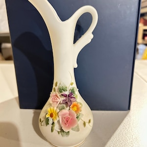 May include: A white porcelain bud vase with a slender neck and a curved handle. The base features a raised, three-dimensional floral arrangement with pink roses, yellow daisies, purple violets, and green leaves. A thin gold band accents the bottom rim.