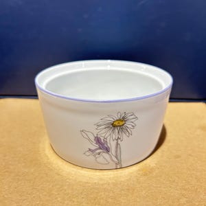 May include: A white ceramic ramekin featuring a delicate botanical illustration of a daisy and an iris on the exterior. The bowl has a smooth, rounded shape with a thin, light purple rim accentuating the top edge, providing a clean and simple look.