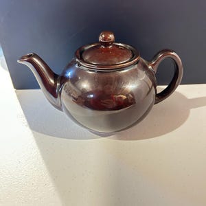 May include: A round, dark brown ceramic teapot with a glossy finish. The teapot features a curved spout, a matching lid with a small knob handle, and a side handle. The surface reflects light, showing a smooth, polished texture throughout the body.