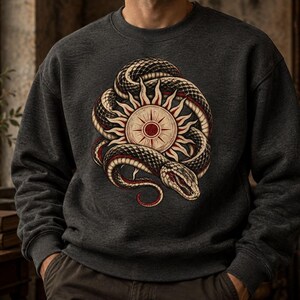 Peut inclure: Un sweat-shirt à col rond gris anthracite foncé arborant une illustration graphique centrale représentant un serpent enroulé autour d'un soleil stylisé aux rayons rayonnants. Le serpent présente des écailles détaillées en noir, blanc et rouge.