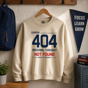 Könnte beinhalten: Ein cremefarbenes Sweatshirt mit Rundhalsausschnitt und dem dunkelblauen Text ERROR 404, ORIGINAL THOUGHT NOT FOUND in Rot sowie BACK TO SCHOOL in Dunkelblau. Das Design enthält horizontale Linien über und unter dem Text auf der Vorderseite.