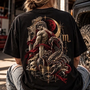 Op de afbeelding: Een zwart t-shirt met een gedetailleerde afbeelding op de achterkant. Het ontwerp toont een gespierd figuur in een rode mantel bij klassieke stenen zuilen, een schedel en een grote schorpioen. Een rode maan en het sterrenbeeld Schorpioen zijn zichtbaar.