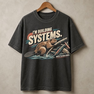 May include: A charcoal grey short-sleeve t-shirt featuring a graphic of a brown beaver building a dam with logs. The text reads I'M BUILDING SYSTEMS. and MOSTLY TO AVOID PEOPLE. The design includes a sunset background with blue water accents.