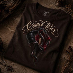 Può includere: Una t-shirt marrone scuro con la grafica di un serpente nero avvolto all'interno di un fiore di calla scuro. Sopra l'illustrazione, il testo "Come Closer" è stampato in un carattere corsivo decorativo di colore chiaro. Scollo rotondo.