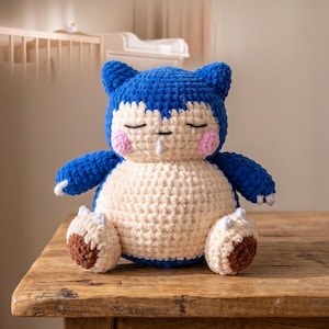 Peut inclure: Un jouet en peluche crocheté à la main représentant une créature bleue et crème avec des oreilles pointues, des yeux fermés, des joues roses et un petit détail blanc près de la bouche. La figurine a des pieds marron foncé et des griffes blanches.