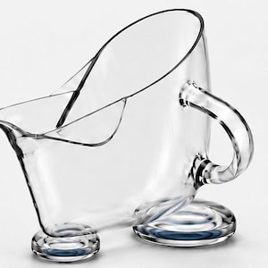 May include: A clear glass gravy boat featuring a wide, curved pouring spout, a sturdy C-shaped handle, and a base supported by three distinct, rounded glass feet. The vessel has a smooth, transparent finish and a wide, open top design.